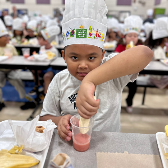 A kid in a chefs hat dunks a banana into a smoothie