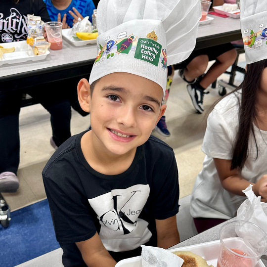 A kid in a chefs hat smiles for the camera
