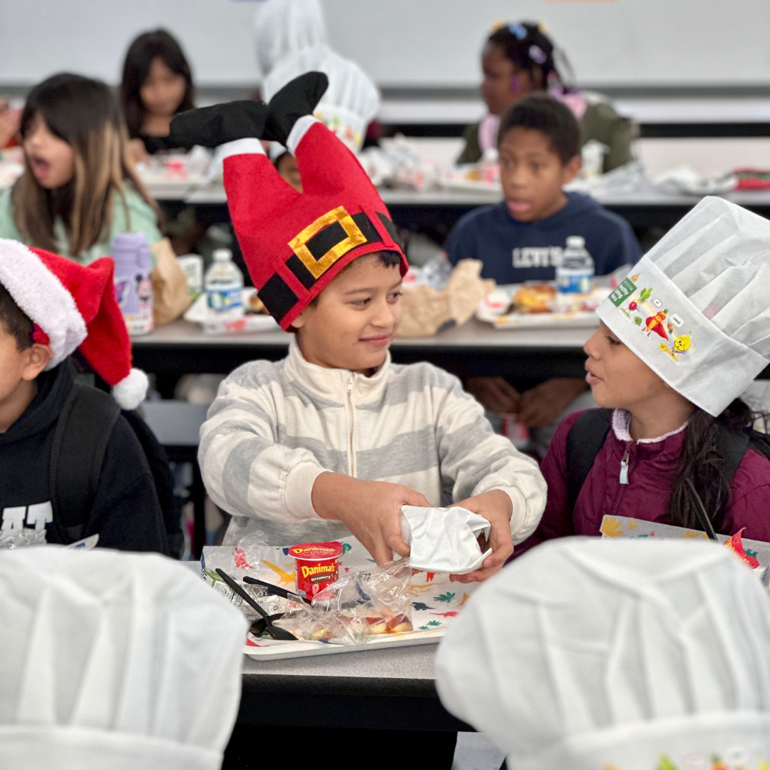 A kid wearing a Santa pats hat and a kid wearing a chefs hat look at each other