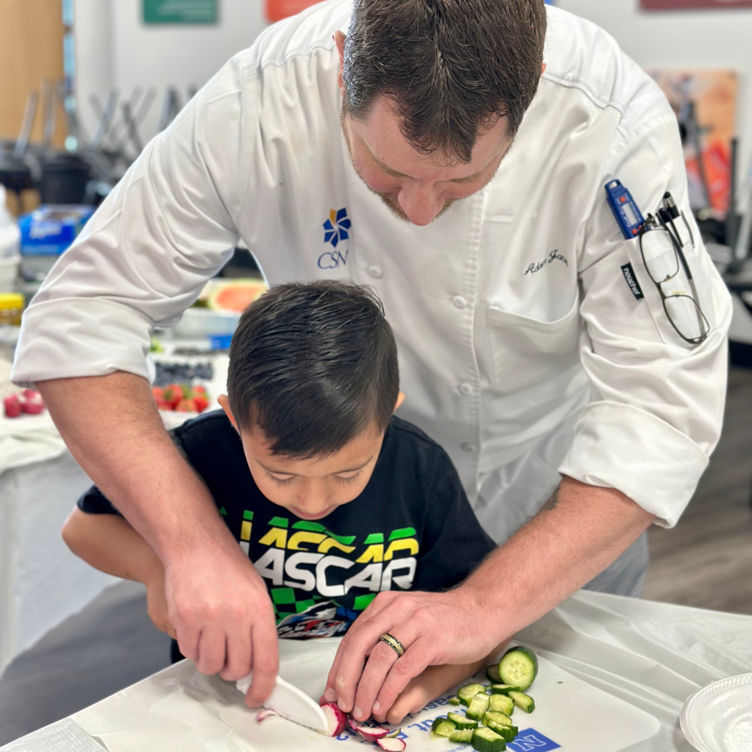 A male chef volunteer teaches a young male student proper knife cutting skills