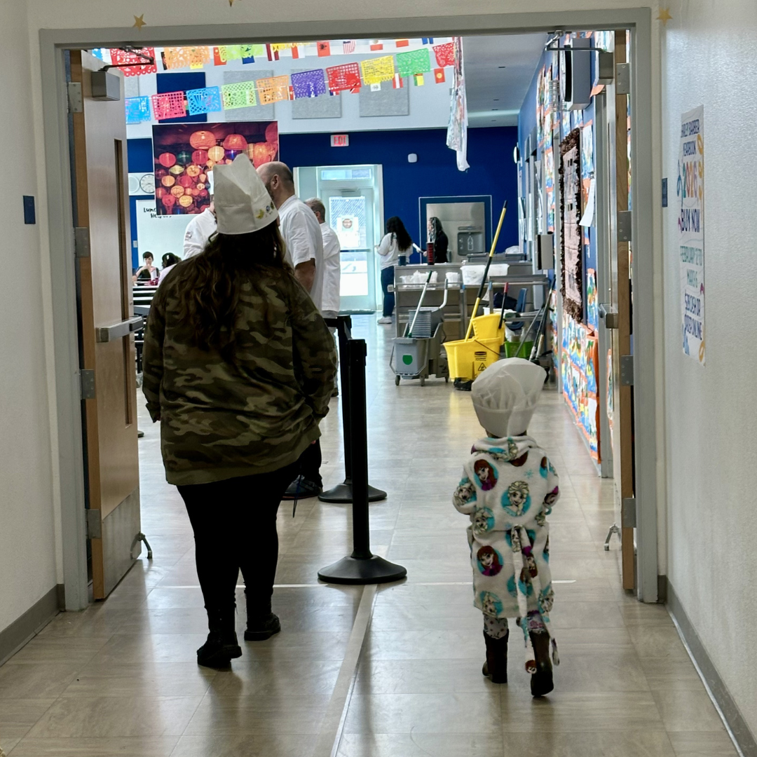 A student and a teacher walking into the cafeteria with chefs hats