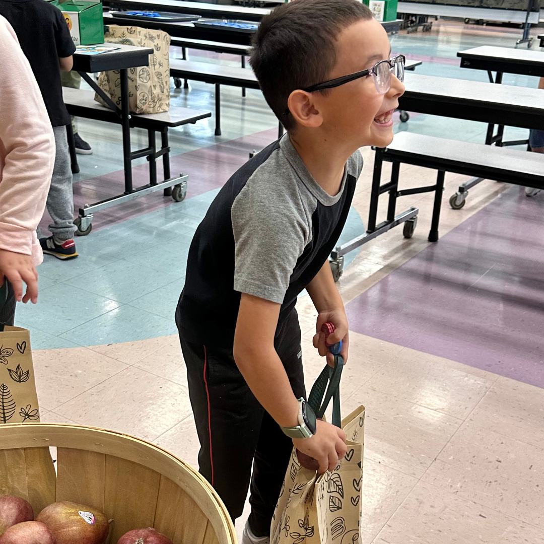 A young male student smiles while putting a red pear in his bag at a farmers market at Ries ES