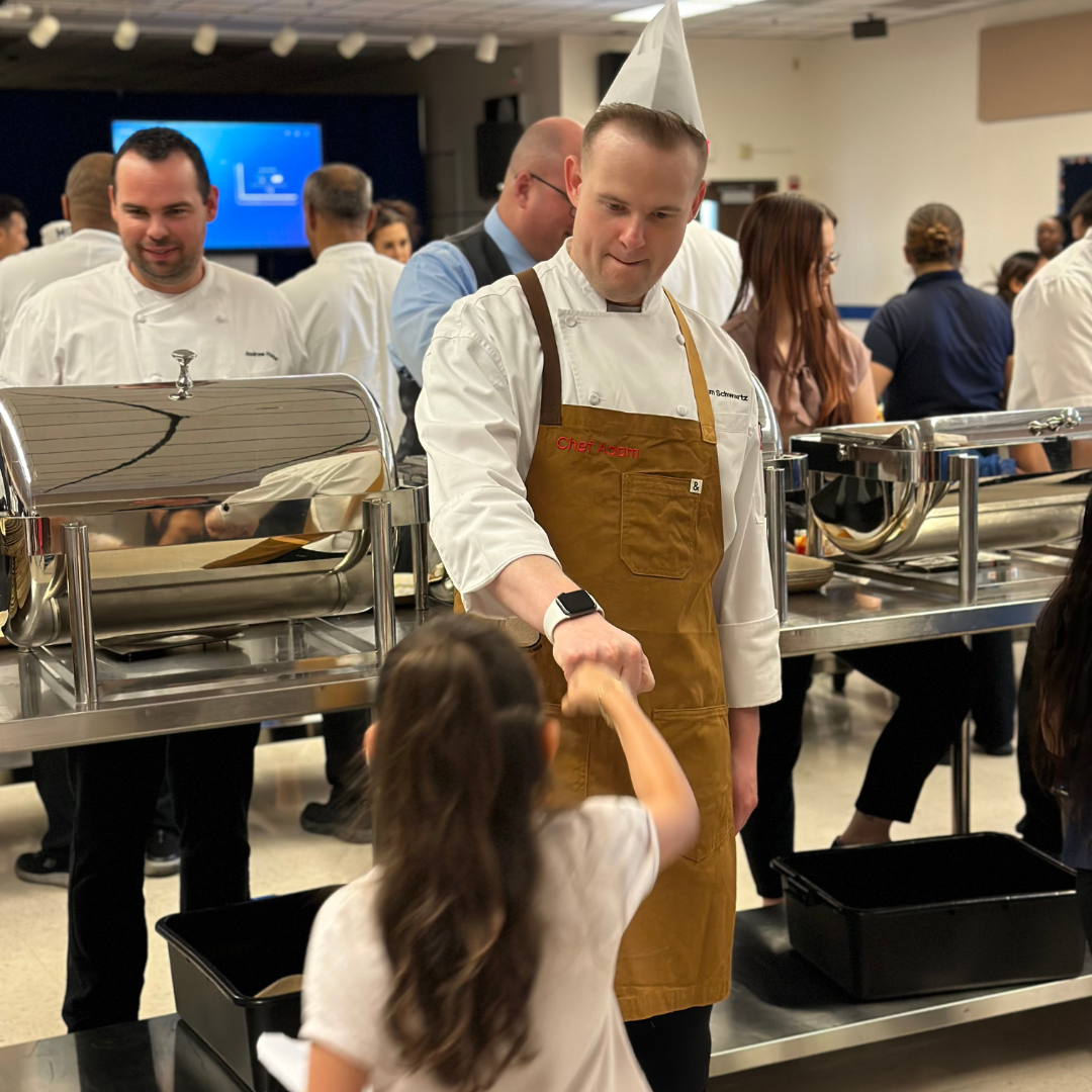 A man in a chefs hate and apron bumps a fist with a kid in a elementary school cafeteria.