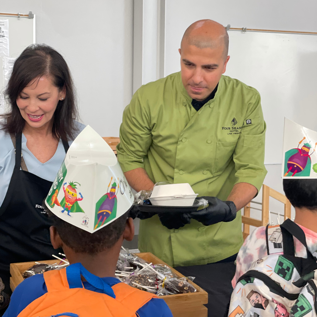 A female and male chef stand behind a table serving plates of food to elementary school students.