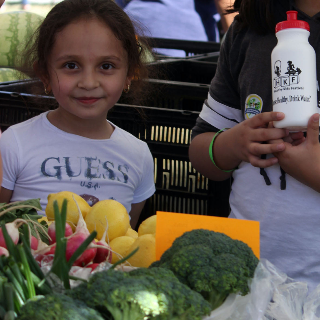 A little girl smiles behind a table of fresh vegetables.
