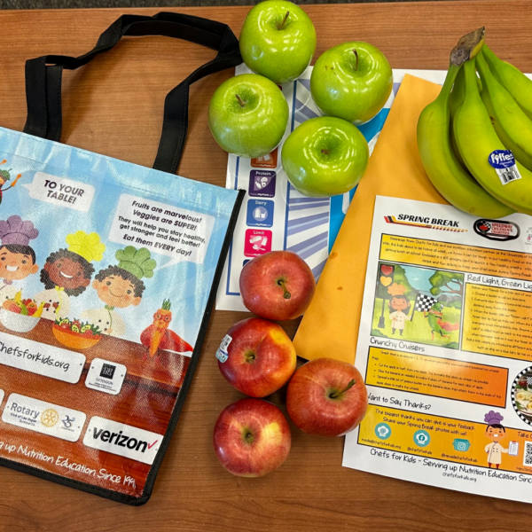 A colorful reusable grocery bag lays on a table next to red and green apples, a bunch of bananas, and some educational flyers.