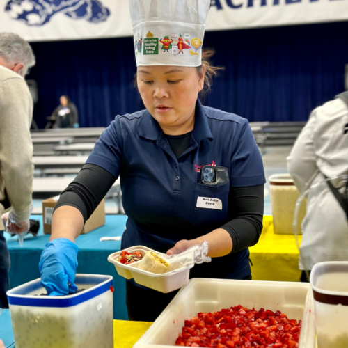 A female chef wearing a chef hat reaches into a bowl of fruit while making an Acai Bowl inside an elementary school cafeteria.