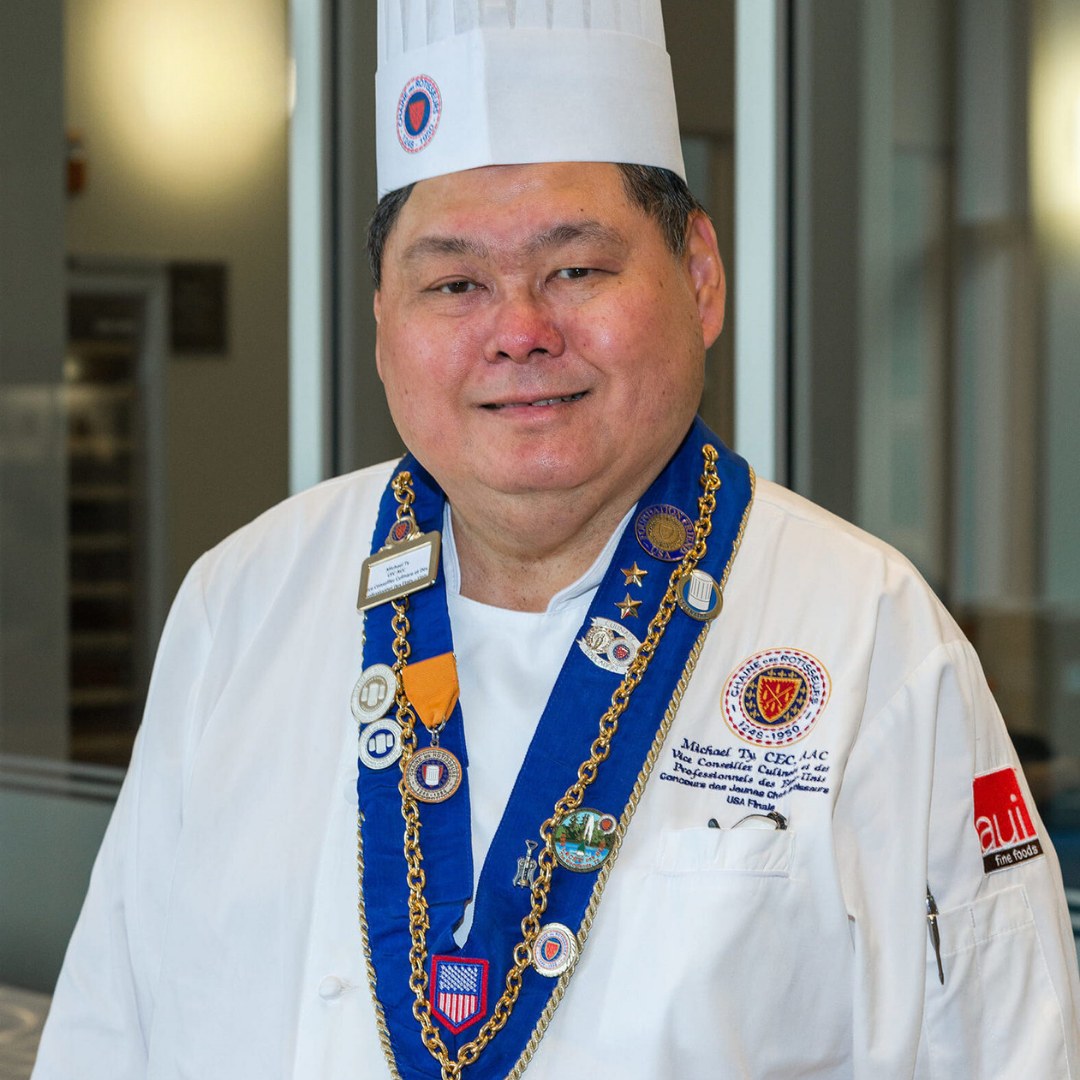 A male chef in a chefs hat and jacket wearing a ribbon around his neck with medals on it.