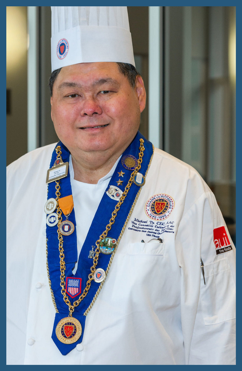 A male chef named Michael Ty poses in a chefs hat & jacket with medals around his neck.