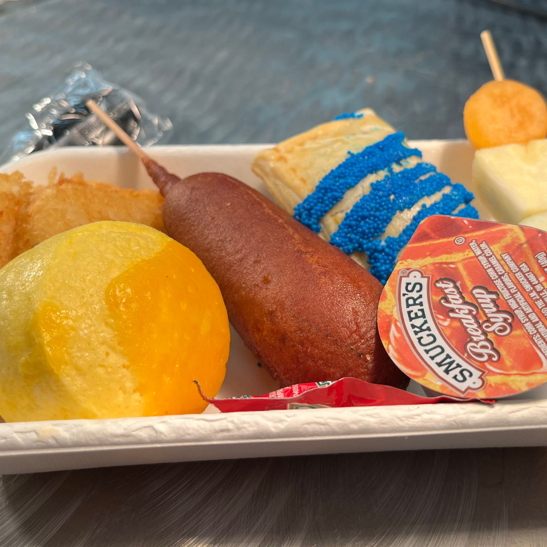 A plate of corn dogs with sausage and pancakes “on a stick,” accompanied by mini cheesy egg bites, hash browns, a fruit skewer, and a scratch-made blueberry poptart in the school colors of Cunningham ES.