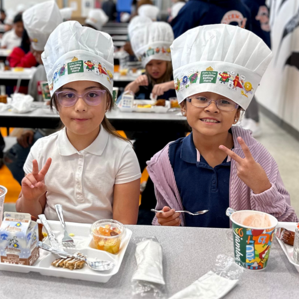 Two young girls wearing chef hats with images of fruits and vegetables dressed as superheroes on them flash the peace sign. They are sitting at a cafeteria table with a plate of food and a smoothie cup in front of them.