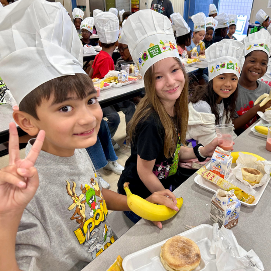 One kid holds of up a peace sign while others are sitting at a breakfast table wearing chefs hats
