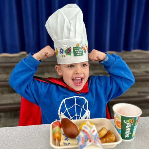 A young kid in a chefs hat, blue shirt, and red cape holds his arms up wit his first close to his head like he's a superhero. He is sitting in front of a breakfast tray.