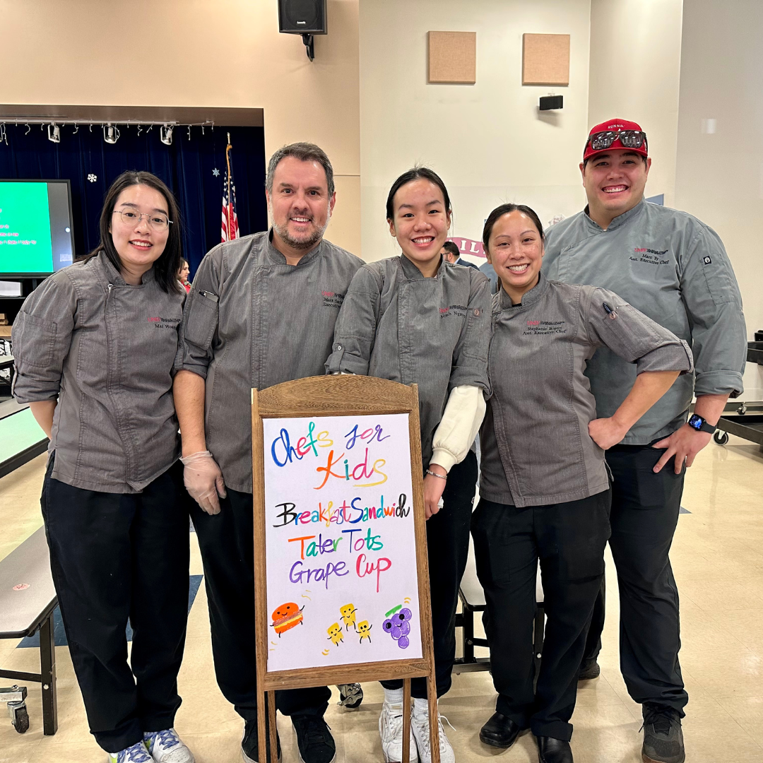 5 people stand in a school cafeteria in front of a sign that reads "Chefs for Kids, Breakfast Sandwich, Tater Tots, Grape Cup." 