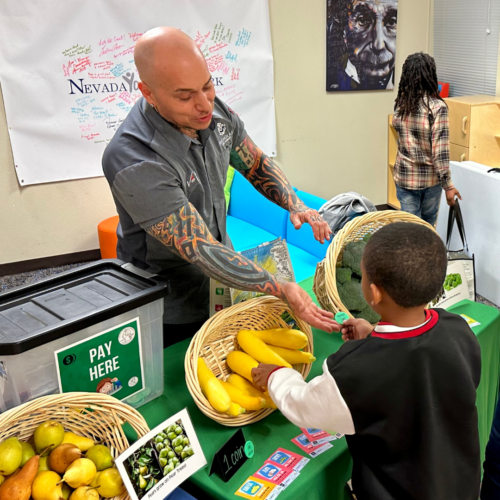 A man and a small child exchange a token for a fresh vegetable at an indoor farmers market.