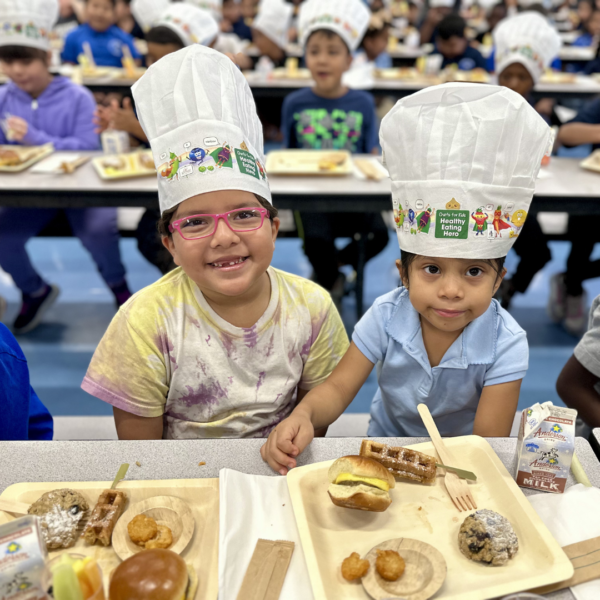 Two female students wearing chef hats smile with trays of food at the breakfast table