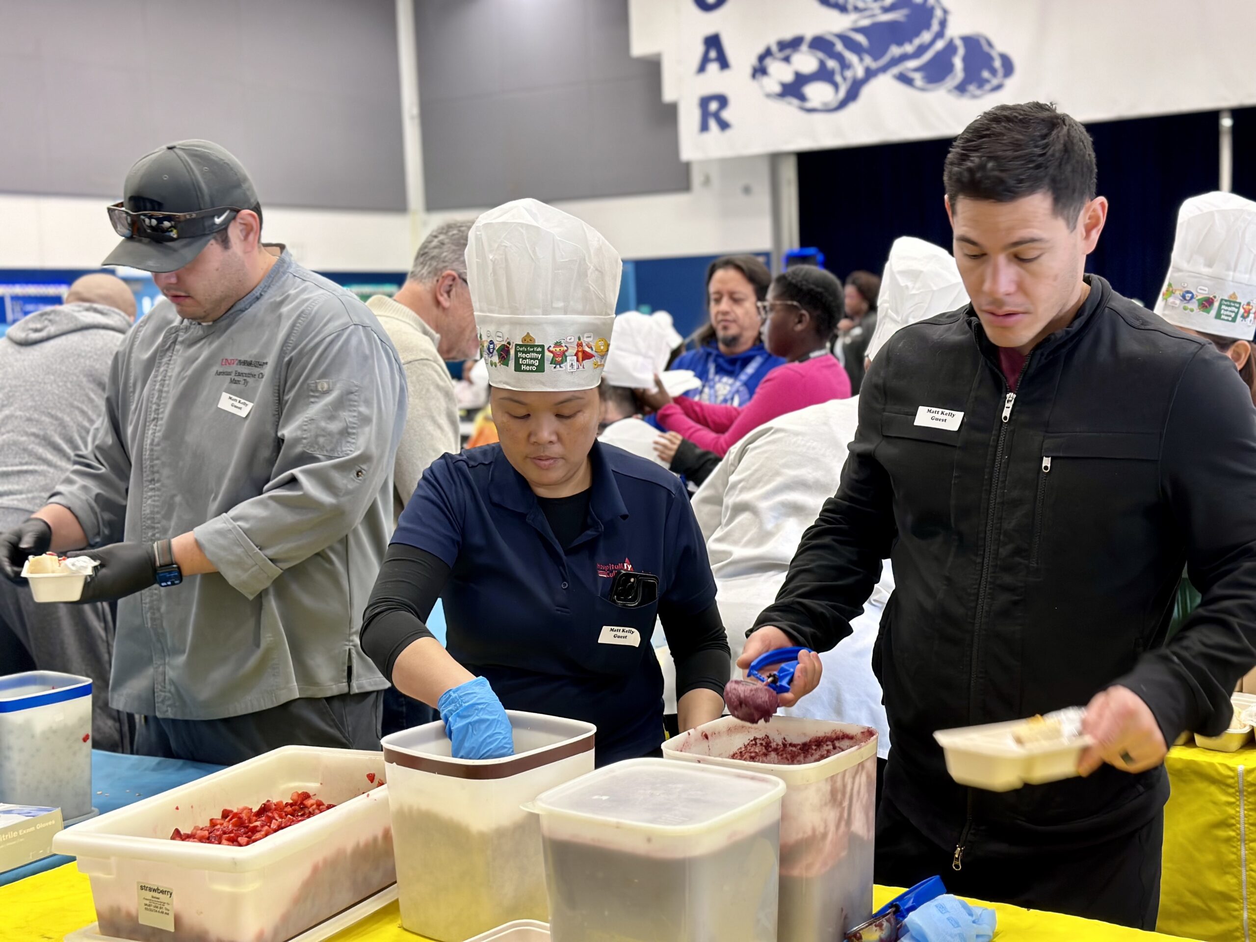 Two men and a female stand behind a table prepping breakfast bowls to be served to kids at an elementary school cafeteria.