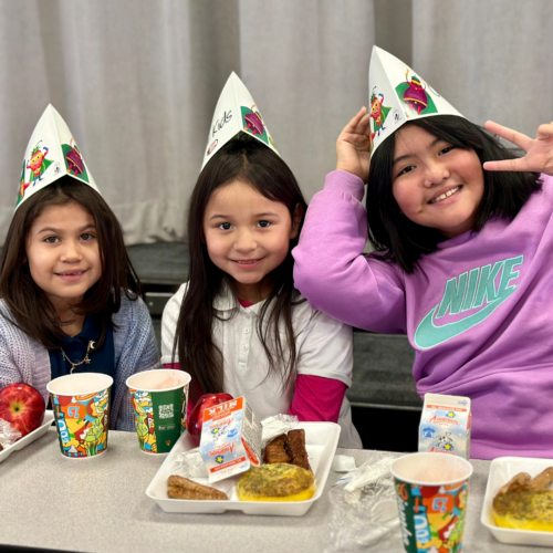 Three girls wearing chef hats pose with lunch trays in a cafeteria.